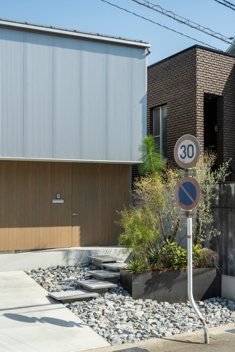 Street view showing corrugated metal and timber panel facades with planted beds along a gravel entrance path