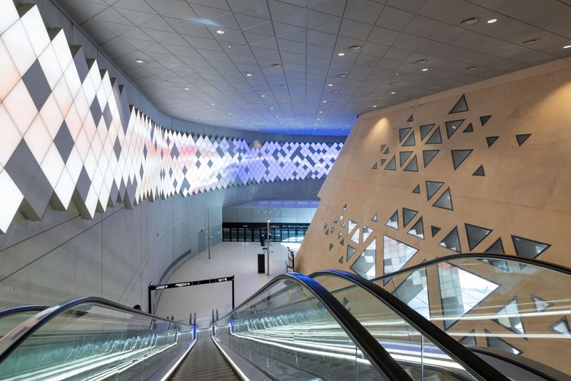 Escalator descending into the station concourse with triangular perforations in concrete walls and geometric ceiling panels