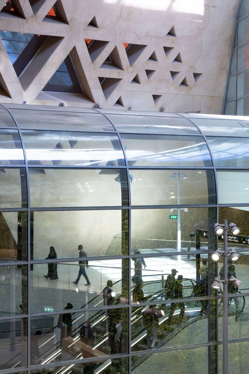 Interior view through curved glass escalator enclosure with concrete wall featuring triangular perforations and visitors descending below