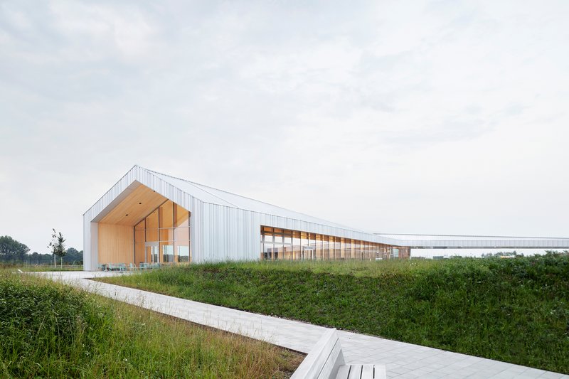 White corrugated metal facade with timber gable entrance beside a concrete walkway through grassy fields