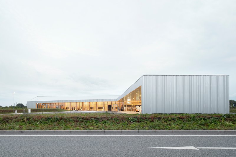 Long metal-clad volume with glazed timber pavilion viewed from the adjacent roadway at dusk