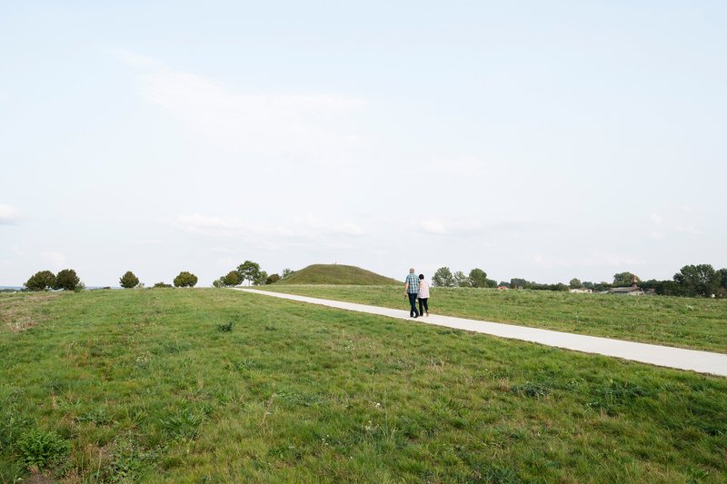 Concrete path crossing a green meadow with two people walking toward a distant mound