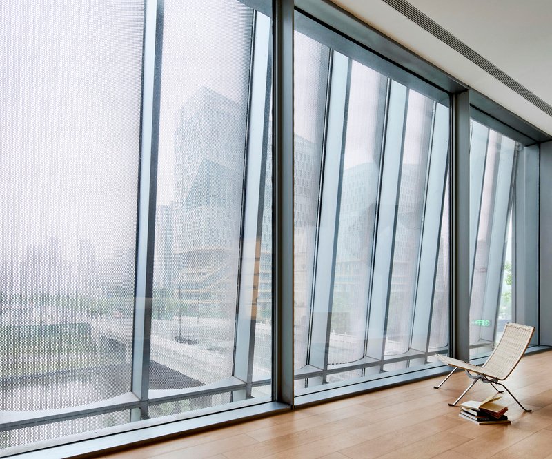 Floor-to-ceiling perforated metal curtain panels filtering daylight over timber flooring with lounge chair