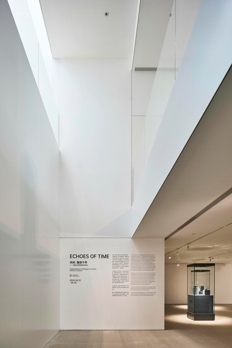 Gallery space with angular white ceiling planes, wall-mounted exhibition text, and illuminated display case