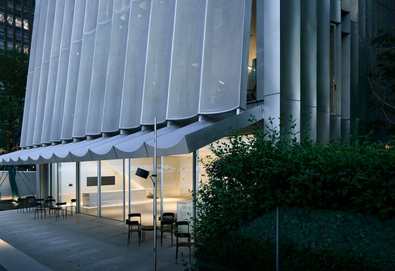 Glass-walled ground floor beneath the scalloped white screen facade at dusk with chairs on the terrace