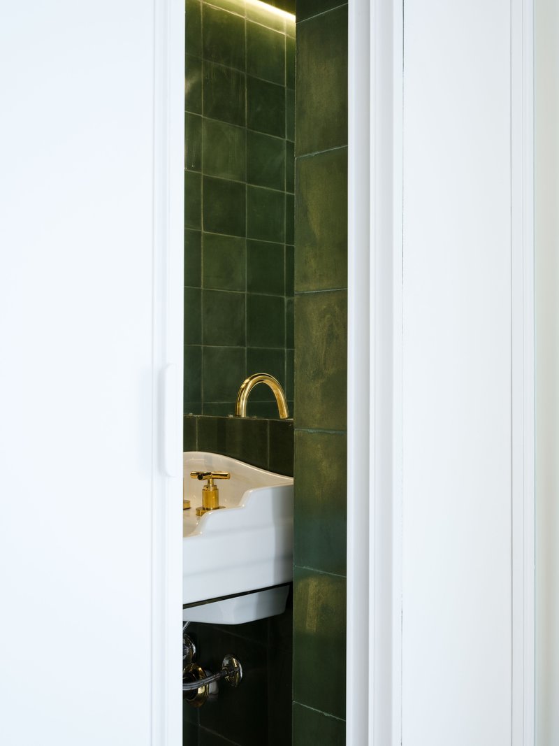 Glimpse through doorway into bathroom with dark green tile walls, brass faucet and concealed strip lighting