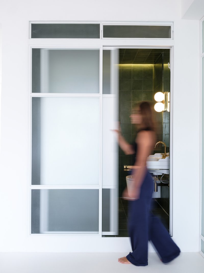 Figure in motion passing through the white-framed sliding door into the olive-tiled bathroom beyond