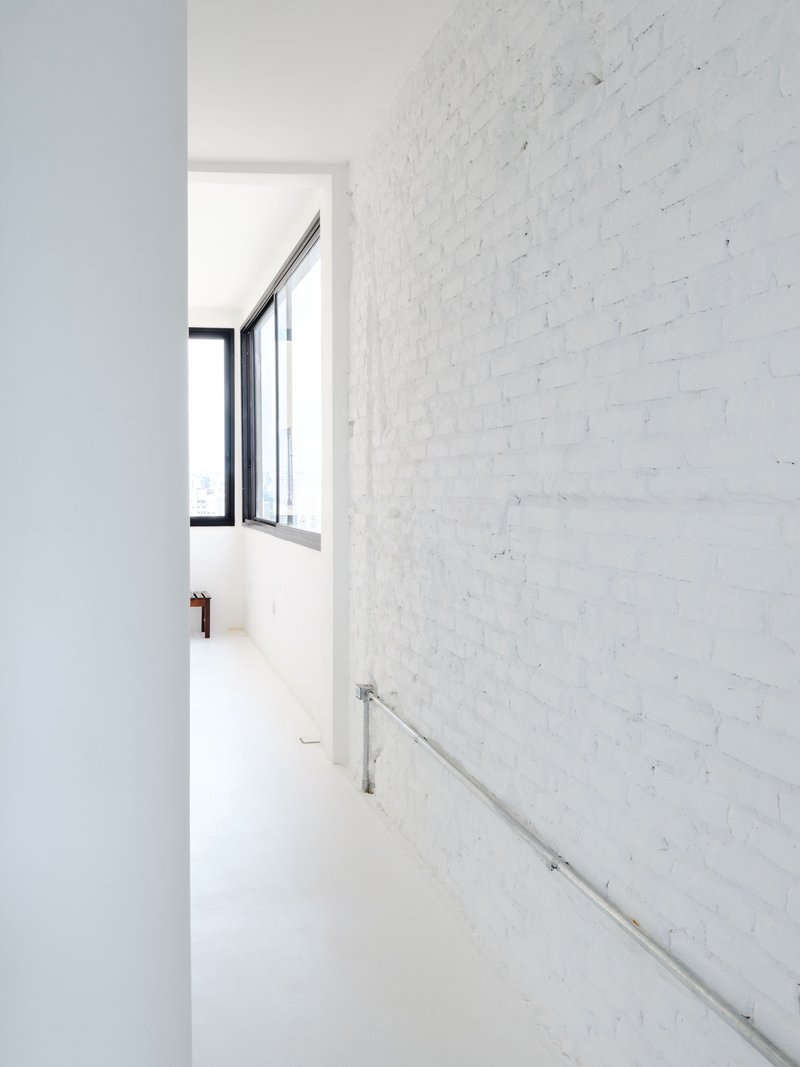 Narrow hallway with white-painted brick wall and stainless steel handrail leading toward a black-framed window