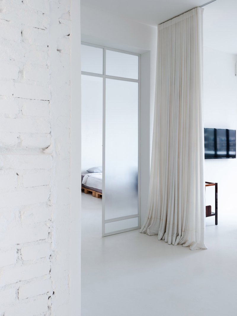 White interior with distressed brick wall, floor-to-ceiling sheer curtains and glimpse of bedroom beyond frosted glass partition