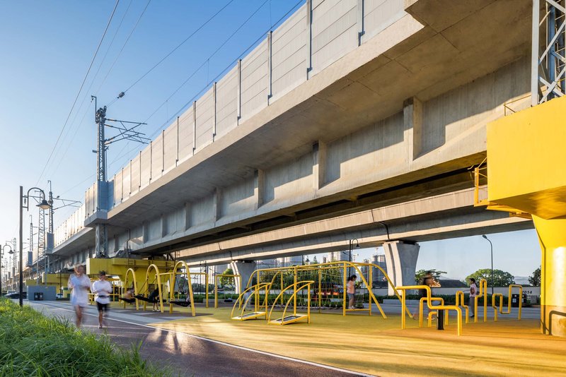 Elevated concrete rail viaduct with yellow playground equipment beneath and pedestrians crossing at dusk