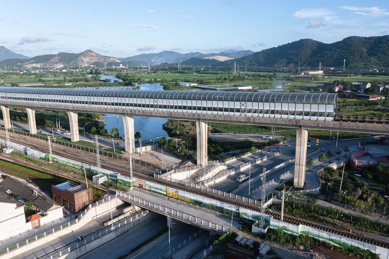Multi-level rail viaduct with arched tubular canopy crossing roadways and green fields below distant mountains