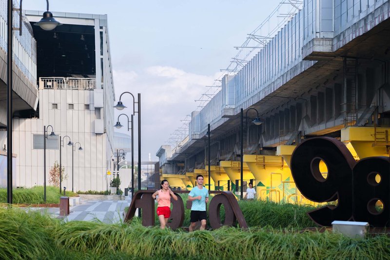 Ground-level plaza with yellow sculptural elements and joggers passing beneath elevated rail infrastructure at dusk