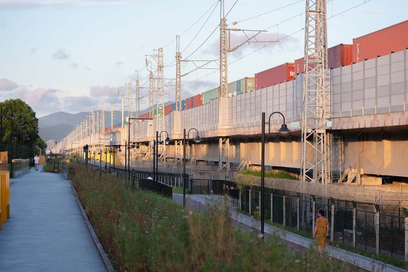 Elevated pedestrian walkway alongside railway infrastructure with corrugated metal walls and overhead catenary towers at dusk