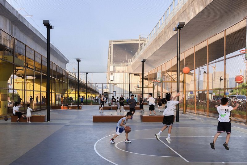 Outdoor basketball court between glass pavilions with players in action and elevated pedestrian bridge above