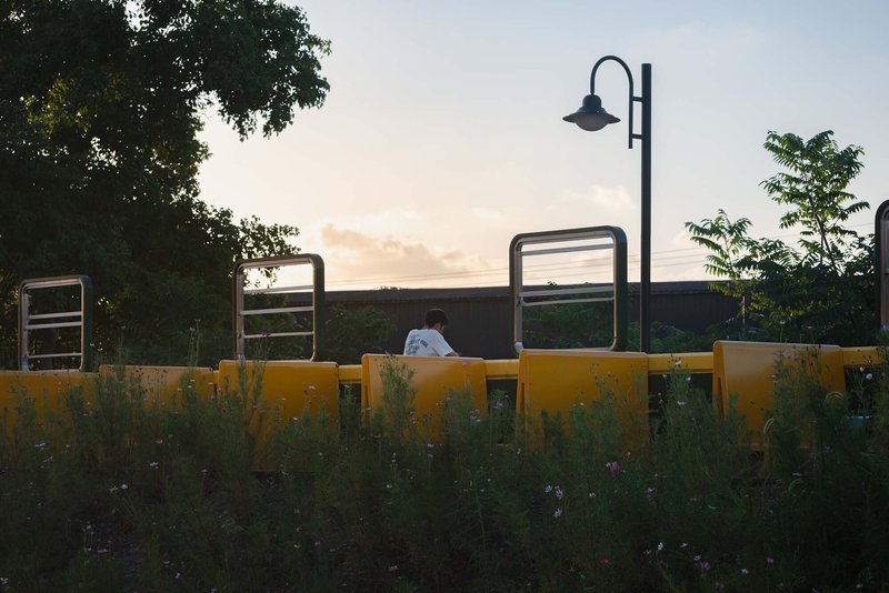 Person seated on yellow bench behind planted buffer with street lamp and exercise equipment at twilight