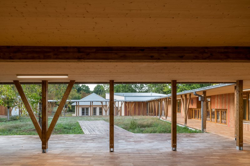 Covered timber walkway with diagonal columns framing views across a planted courtyard to adjacent timber structures