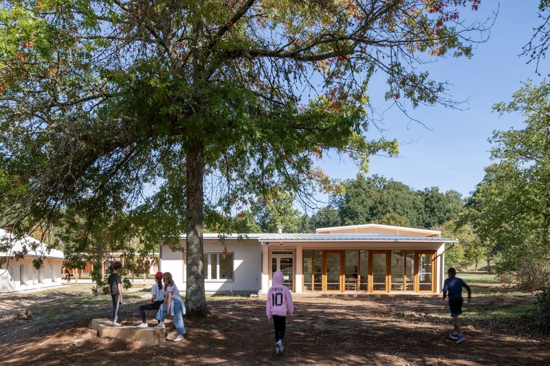 Low pavilion with metal roof and glazed timber facade as children gather beneath a tree