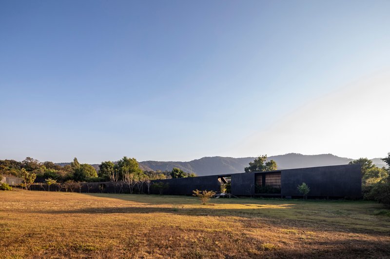 Long concrete facade with small window openings facing a dry grass field at dusk