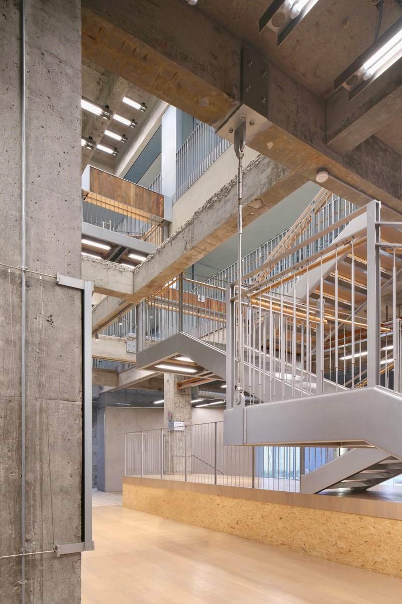 Multi-level interior atrium with exposed concrete columns, timber beams, and metal staircases under fluorescent lighting