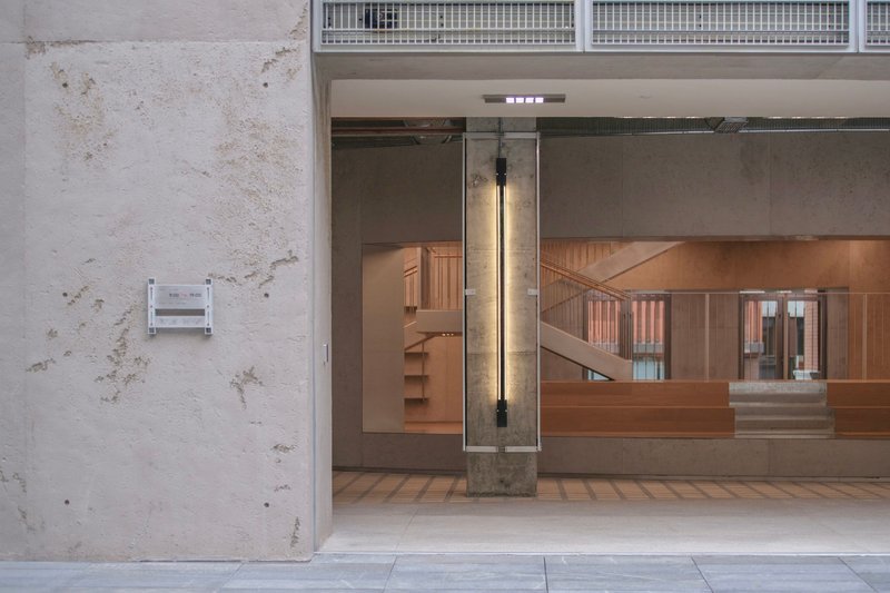View through concrete portal showing timber volume with metal railing and exposed cylindrical column with wall-mounted light