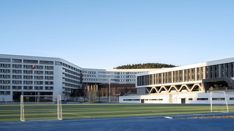 View across the athletic field showing zigzag canopy structure connecting white residential wings and hillside backdrop