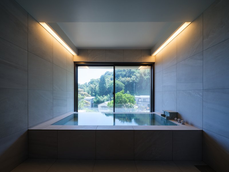 Soaking tub with stone tile surround and recessed ceiling lighting facing hillside landscape
