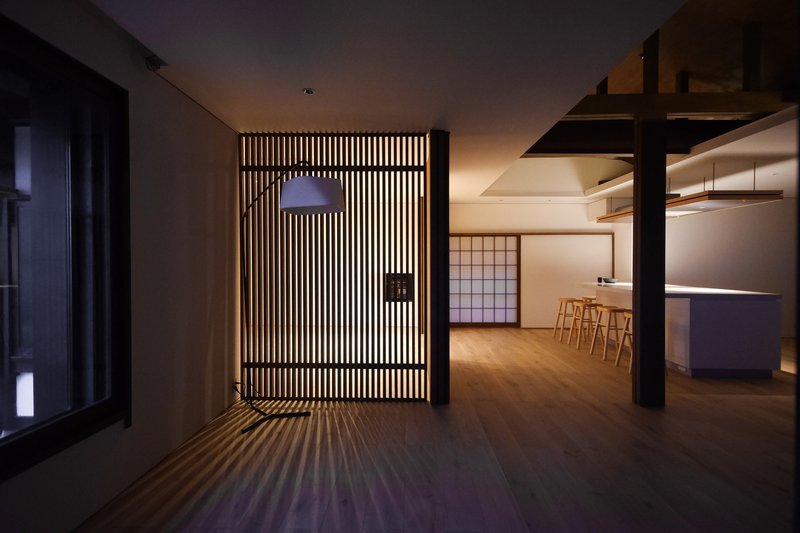 Vertical timber slat screen with backlit striping casting shadow patterns across the floor at dusk