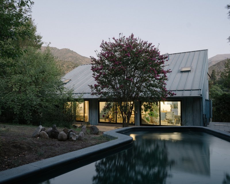 Gabled metal roof with glazed walls framing a tree at dusk, reflected in a dark pool