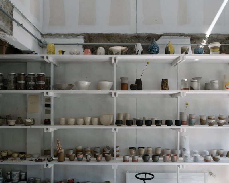 White metal shelving filled with ceramic vessels and pottery beneath a raw concrete beam