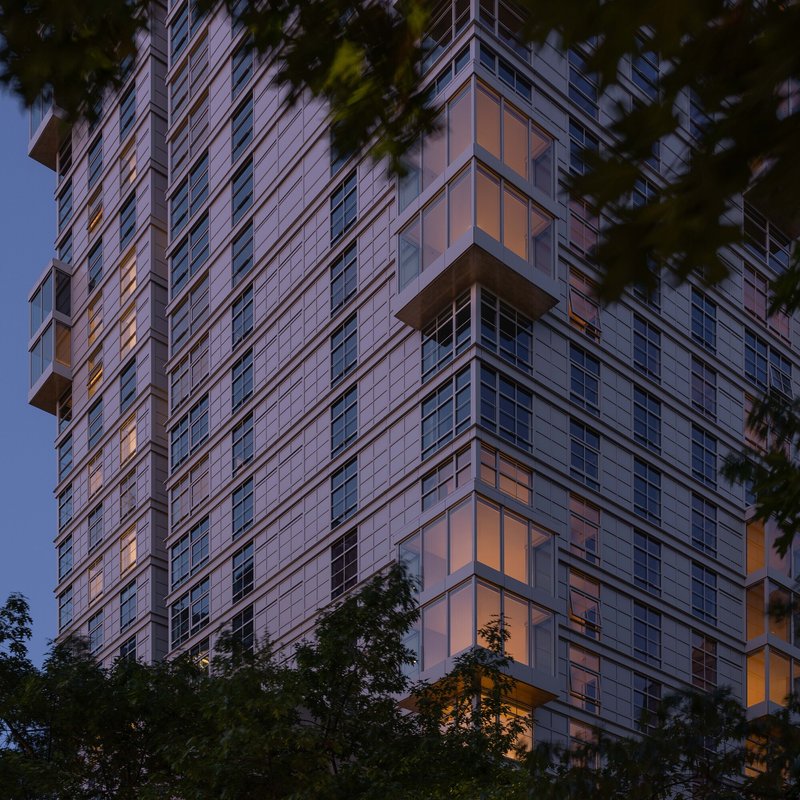 Glass and panel facade of a residential tower framed by trees at dusk with interior lights glowing