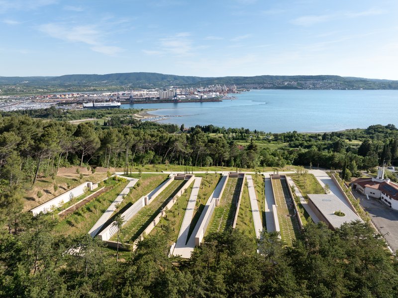 Aerial view of parallel green-roofed pavilions arranged across a hillside overlooking a bay and distant mountains