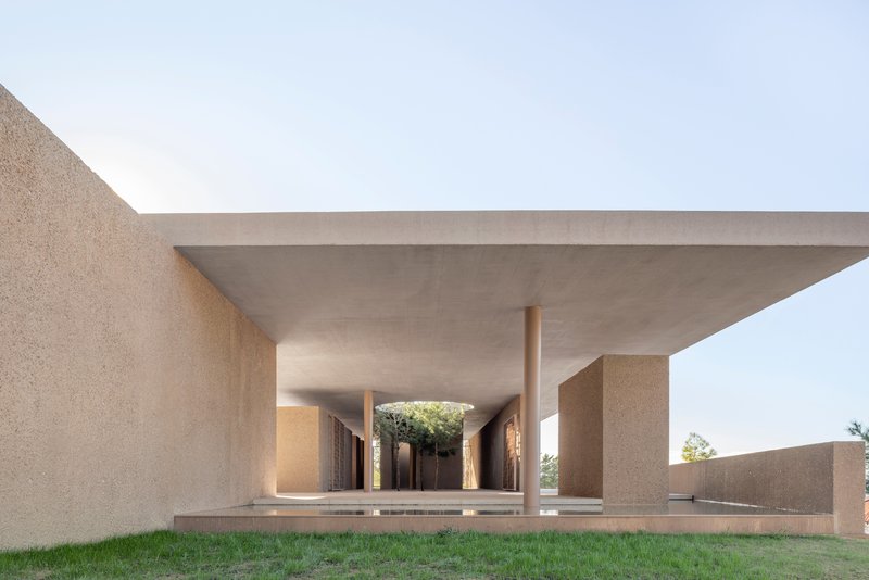 Covered colonnade with cylindrical columns supporting a concrete slab roof above a lawn in afternoon light