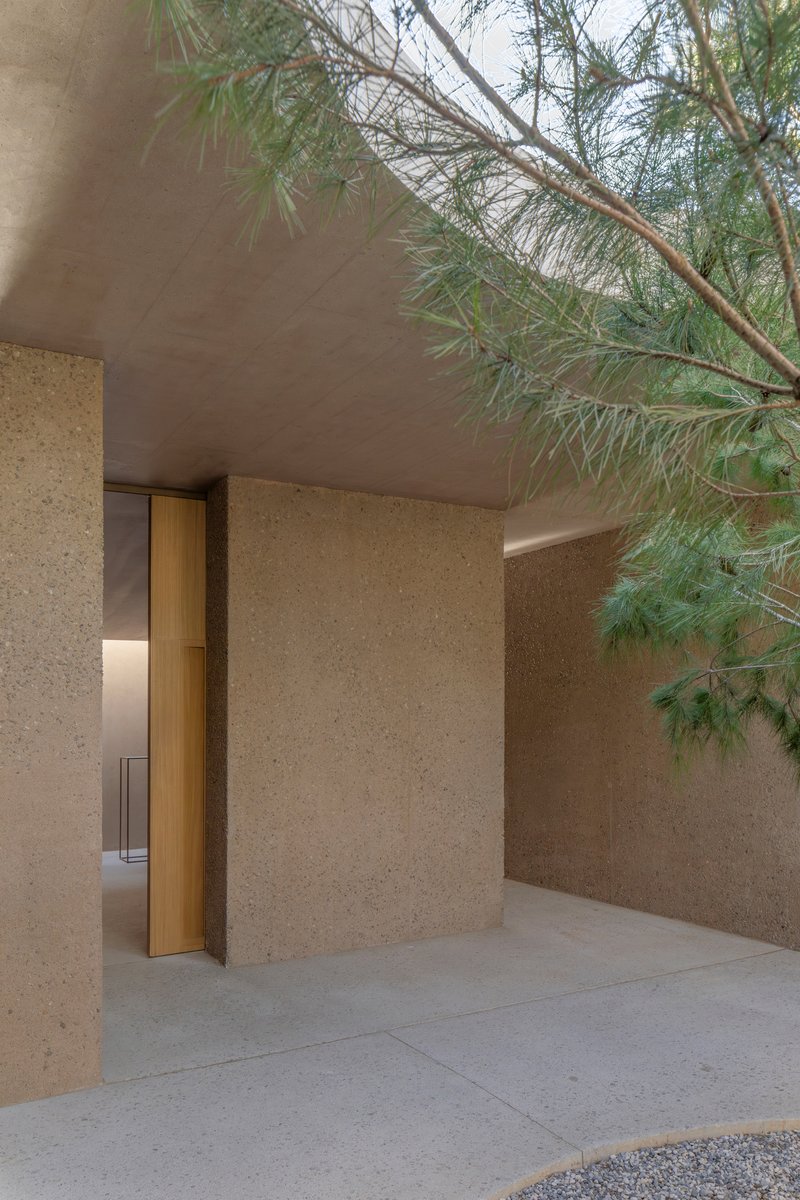 Recessed timber doorway in a textured aggregate wall beneath pine branches and an overhanging soffit