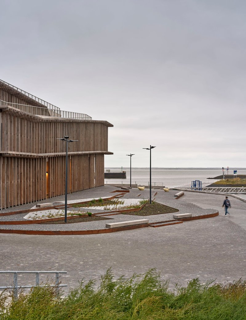 View of the curved timber facade with terraced paving and a lone figure walking along the waterfront