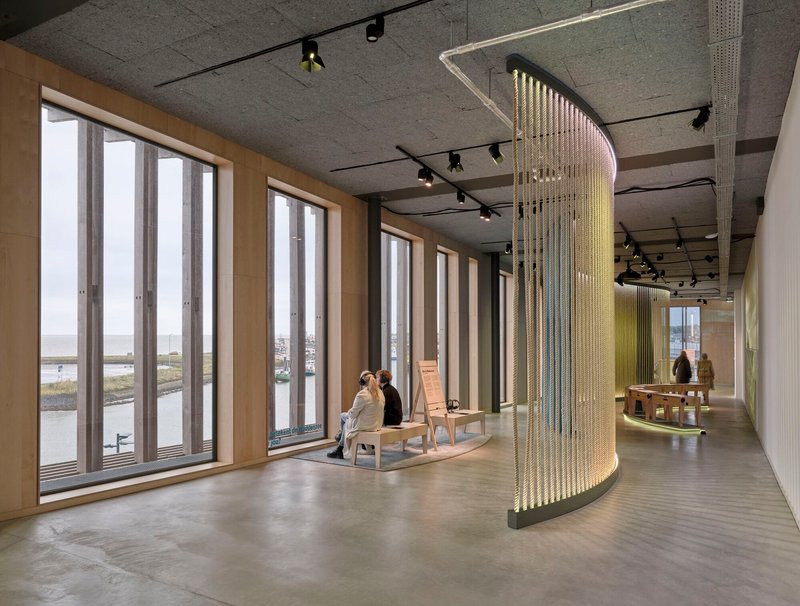 Curved brass bead curtain installation beside tall timber-framed windows with person seated near waterfront view