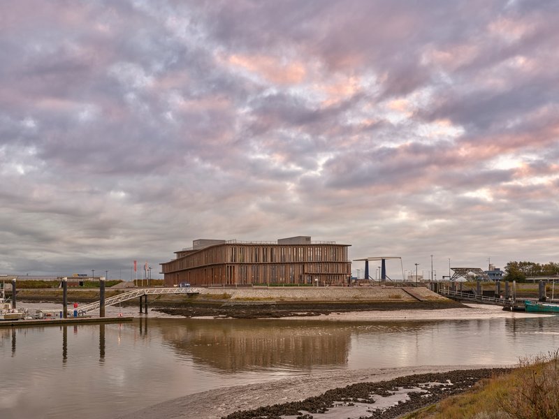 Horizontal timber-clad building reflected in the tidal waters of a mudflat harbor under dramatic clouds