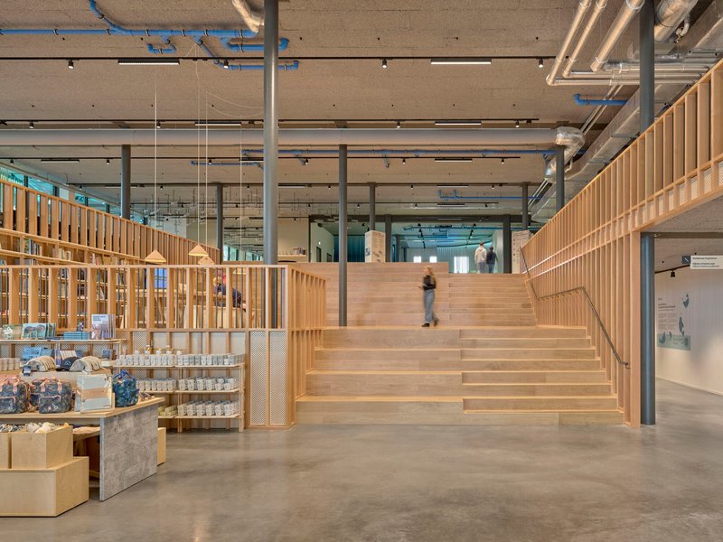 Wide timber staircase ascending past a retail display wall under an exposed concrete soffit with mechanical ducts