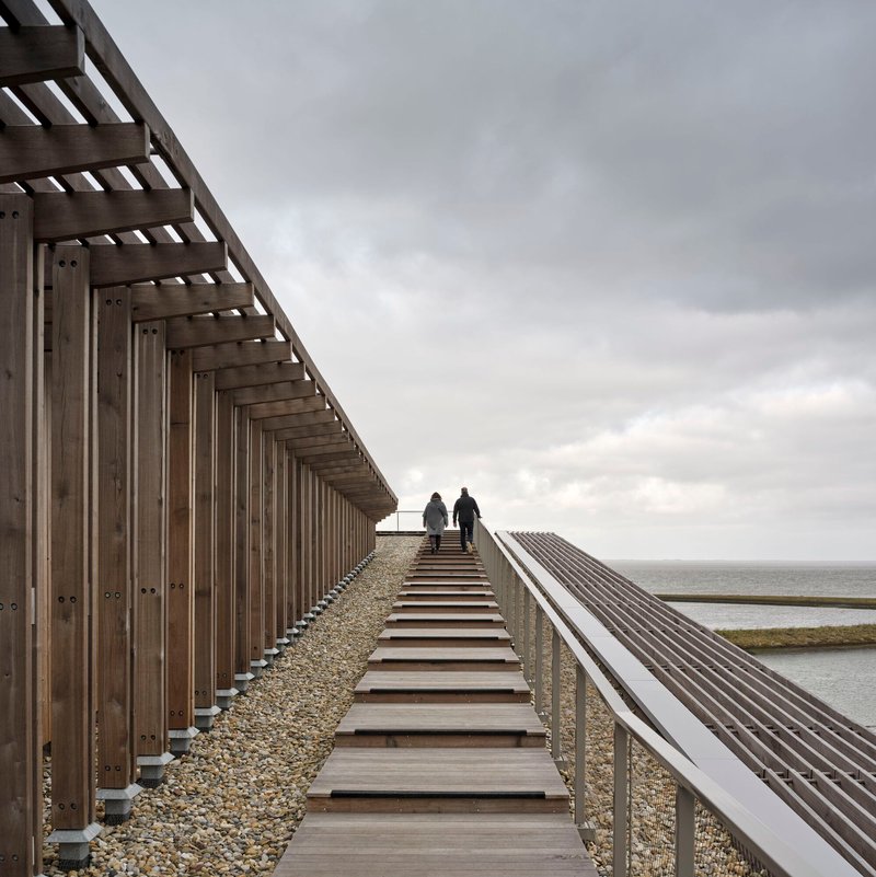 Timber boardwalk with open stairway alongside vertical slat screen and gravel ballast roof overlooking the sea