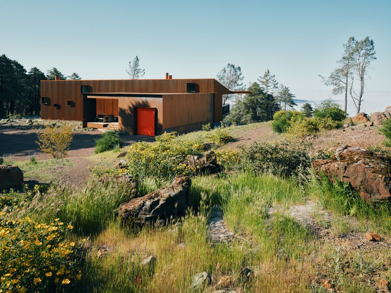 Weathered steel and timber residence with red entry door set among wildflowers and volcanic rock outcroppings
