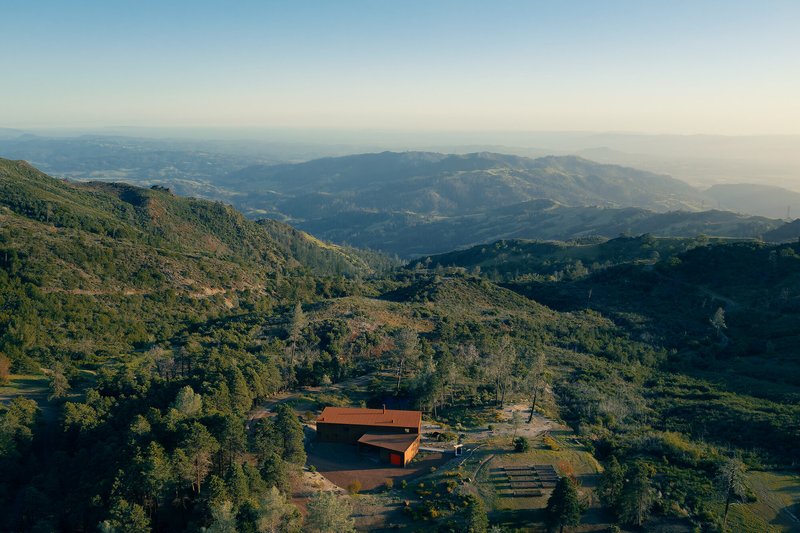 Aerial view of weathered steel-clad buildings on terraced hillside amid forested mountain terrain