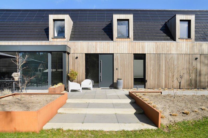 Vertical timber facade with projecting dormer windows above a concrete paver path and planted steel beds