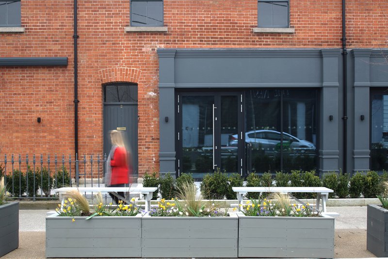 Red brick facade with grey metal-framed storefront and painted planter boxes with yellow flowers and grasses