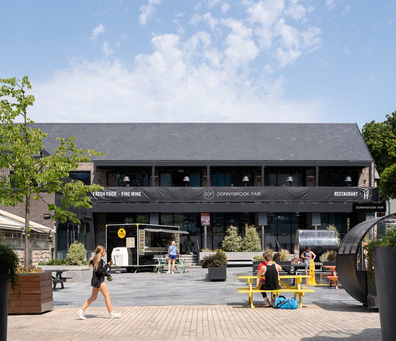 Public plaza with yellow picnic tables and pedestrians in front of two-story commercial building