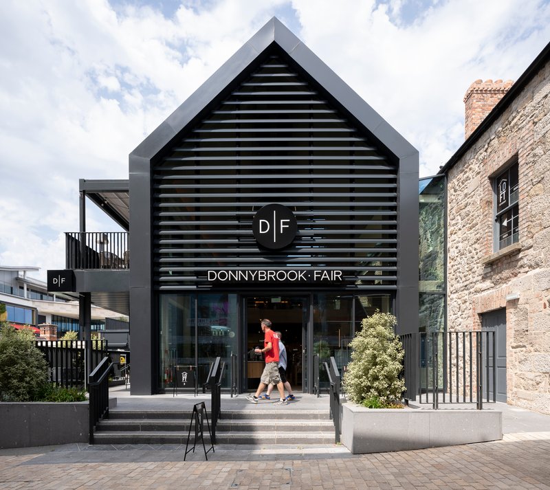 Gable-fronted entrance with horizontal black louvers and pedestrians ascending concrete steps under cloudy sky