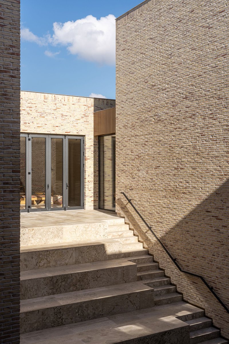 Stone staircase ascending between buff brick walls toward glazed doors beneath blue sky