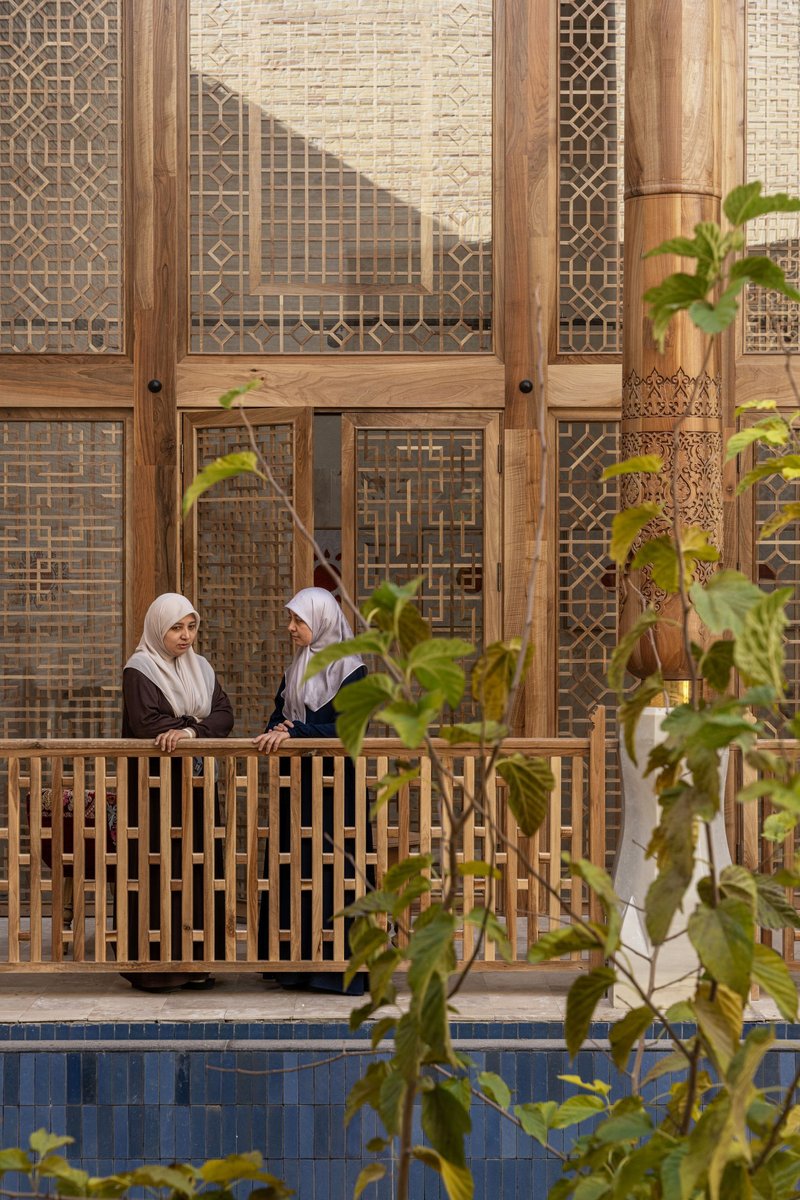 Timber lattice balcony overlooking blue-tiled pool with two visitors and planted foliage in foreground