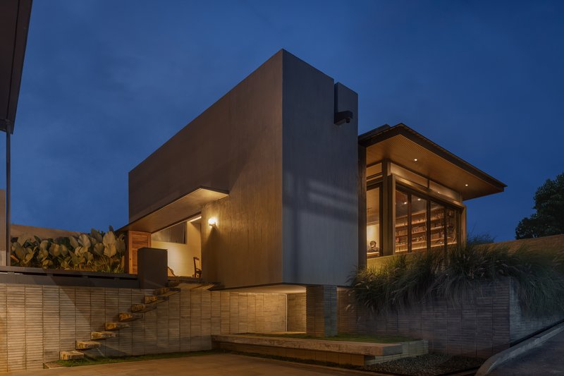 Street-facing facade with exposed concrete tower, external stair alongside a tile-clad retaining wall at twilight