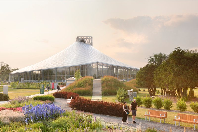 White tensile roof rising to a central lantern above a glass pavilion surrounded by planted beds with visitors