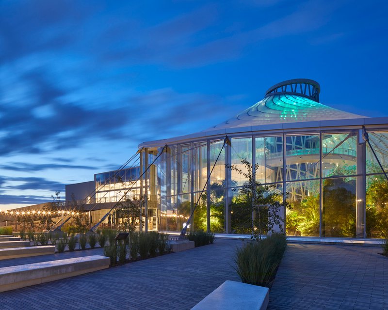 Glass conservatory volume with exposed steel structure and central dome illuminated against a blue evening sky