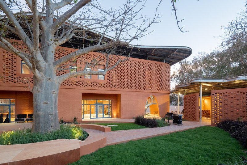 Perforated terracotta facade with curved volumes surrounding a courtyard garden with mature baobab tree at dusk